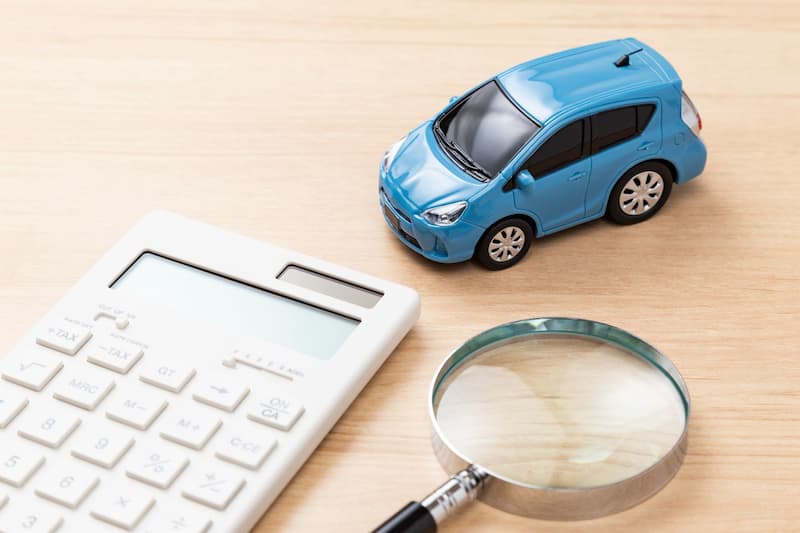デスク上の青いミニカーと電卓と虫眼鏡が示す自動車費用の安心価格のイメージ