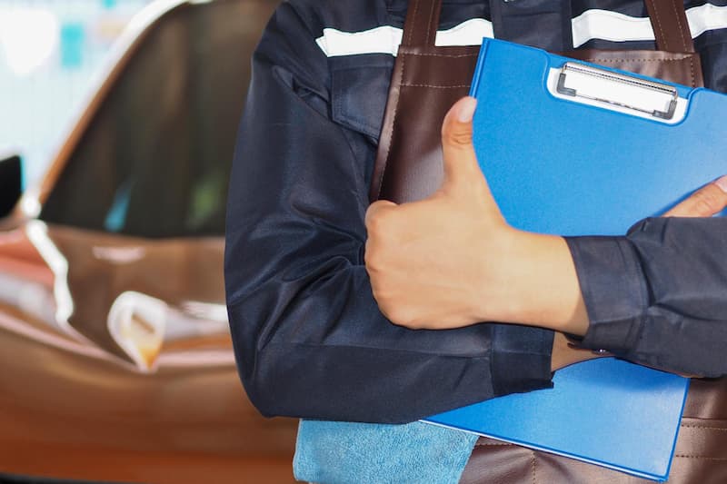 青いクリップボードを持つ整備士が車の点検作業をしている様子