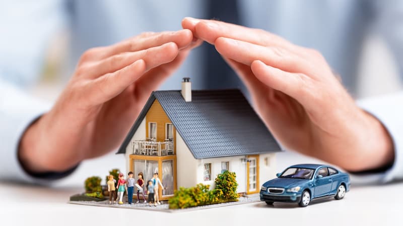 両手で守られている家族のミニチュア住宅と車を保険で保護する日常のドライバーのイメージ